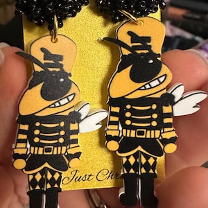 May include: A pair of novelty earrings featuring a cartoon character in yellow, black, and white. The earrings have black beaded tops and are hanging from a gold-colored hook. The character has a smiling face and is wearing a uniform.