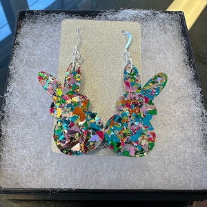 Może przedstawiać: Para srebrnych kolczyk&oacute;w wiszących z kolorowym, błyszczącym wzorem kr&oacute;lika. Kr&oacute;lik jest wykonany z tęczowego, wielokolorowego brokatu i ma efekt 3D.