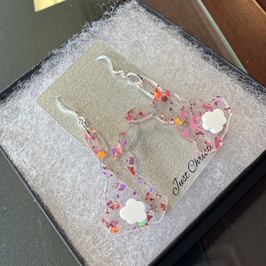 May include: A pair of pink and iridescent glitter bunny earrings with white cloud charms. The earrings are displayed in a black box with white tissue paper. The box has the text "Just Christie" printed on it.