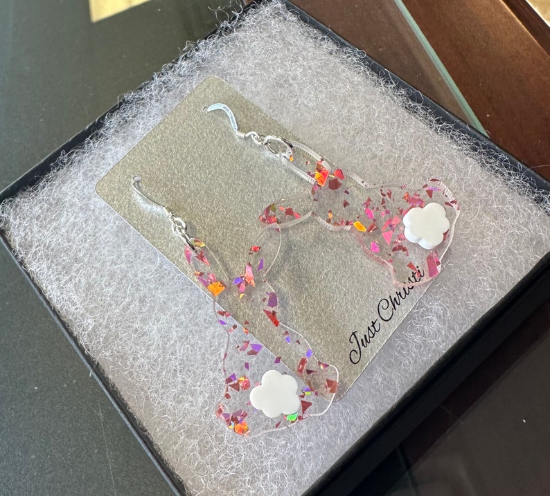 May include: A pair of pink and iridescent glitter bunny earrings with white cloud charms. The earrings are displayed in a black box with white tissue paper. The box has the text "Just Christie" printed on it.