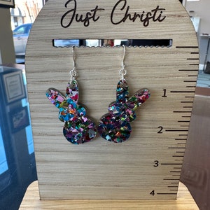 Może przedstawiać: Para kolorowych błyszczących kolczyk&oacute;w w kształcie kr&oacute;liczych uszu na drewnianym stojaku z wygrawerowanym tekstem "Just Christi" na g&oacute;rze.