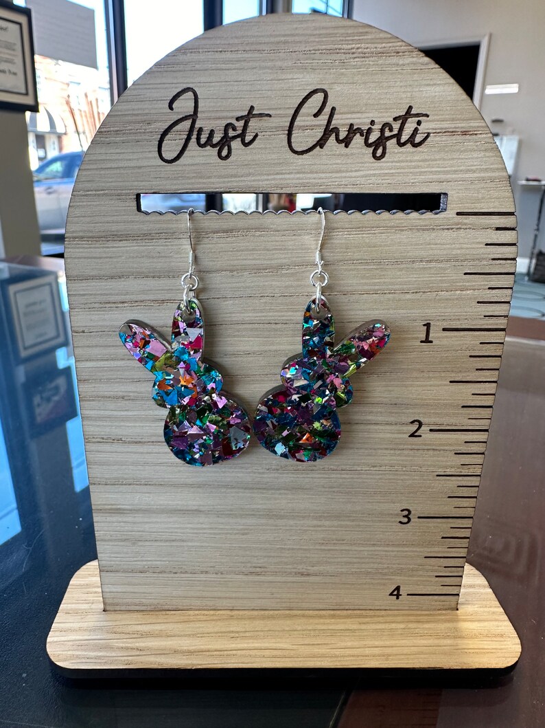 Może przedstawiać: Para kolorowych błyszczących kolczyk&oacute;w w kształcie kr&oacute;liczych uszu na drewnianym stojaku z wygrawerowanym tekstem "Just Christi" na g&oacute;rze.