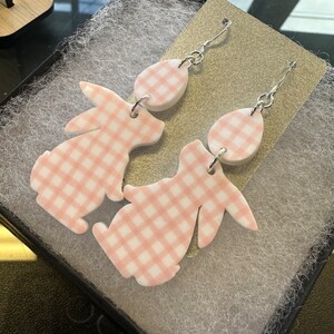 Puede incluir: Un par de pendientes de conejito de gingham rosa y blanco con ganchos plateados. Los pendientes est&aacute;n hechos de arcilla polim&eacute;rica y tienen forma de conejitos con patrones de gingham.
