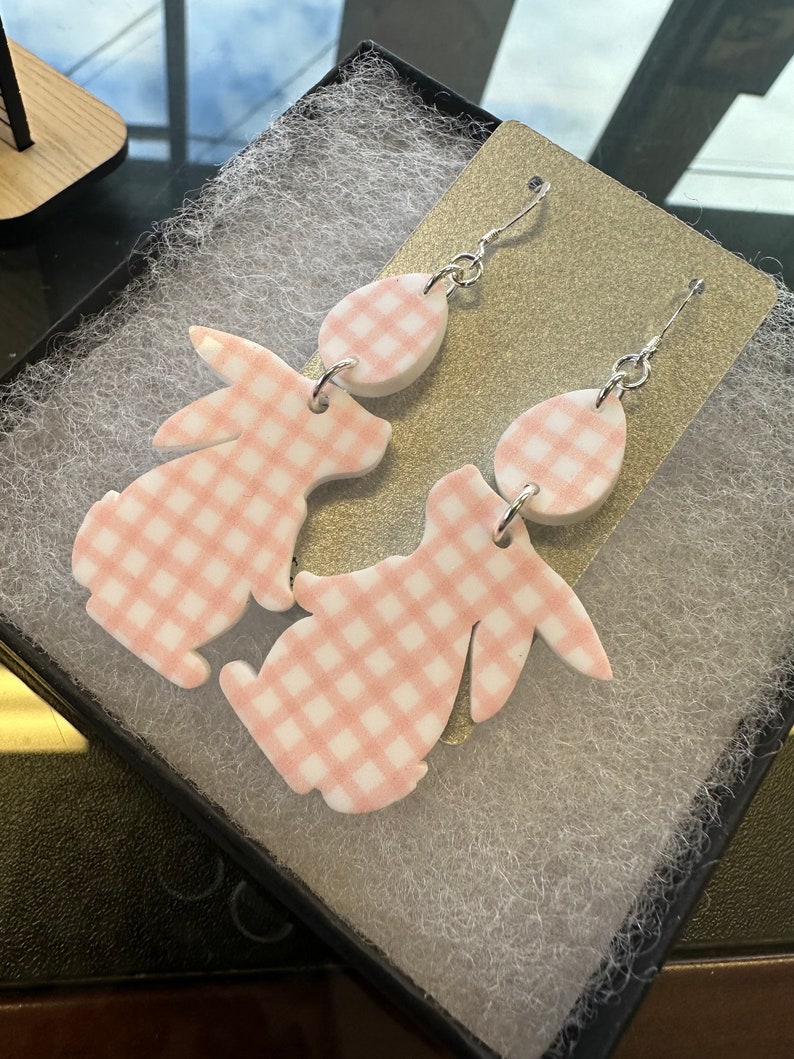 Puede incluir: Un par de pendientes de conejito de gingham rosa y blanco con ganchos plateados. Los pendientes est&aacute;n hechos de arcilla polim&eacute;rica y tienen forma de conejitos con patrones de gingham.