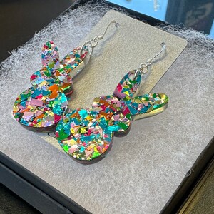 Może przedstawiać: Para srebrnych kolczyk&oacute;w wiszących z kolorowym, błyszczącym wzorem kr&oacute;lika. Kr&oacute;lik jest wykonany z żywicy i pokryty małymi, tęczowymi kawałkami brokatu.