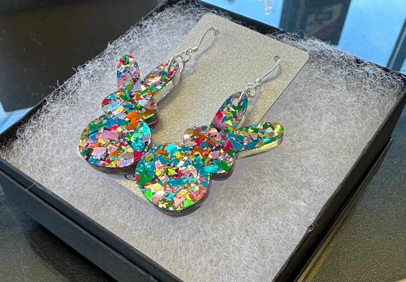 Może przedstawiać: Para srebrnych kolczyk&oacute;w wiszących z kolorowym, błyszczącym wzorem kr&oacute;lika. Kr&oacute;lik jest wykonany z żywicy i pokryty małymi, tęczowymi kawałkami brokatu.