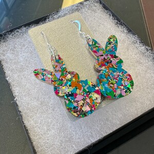 Może przedstawiać: Para kolczyk&oacute;w wiszących z kolorową sylwetką kr&oacute;lika. Kr&oacute;lik jest wykonany z błyszczącego materiału w połączeniu kolor&oacute;w r&oacute;żowego, niebieskiego, zielonego i pomarańczowego.