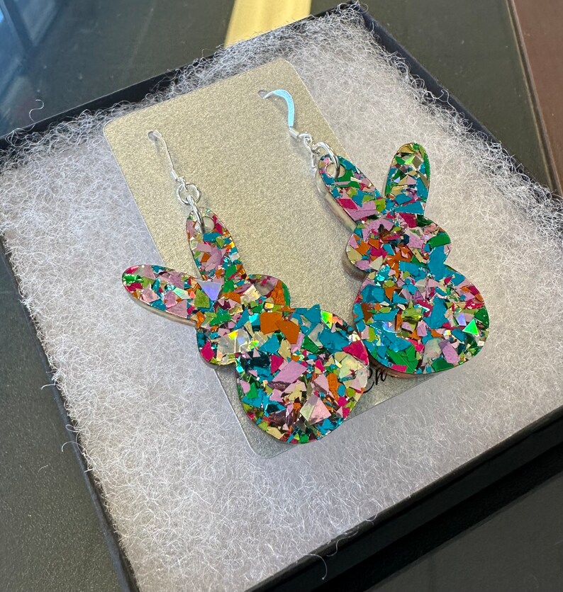 Może przedstawiać: Para kolczyk&oacute;w wiszących z kolorową sylwetką kr&oacute;lika. Kr&oacute;lik jest wykonany z błyszczącego materiału w połączeniu kolor&oacute;w r&oacute;żowego, niebieskiego, zielonego i pomarańczowego.