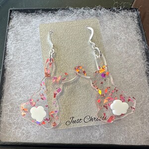 May include: A pair of pink and orange glitter bunny rabbit earrings with white accents. The earrings are displayed in a white box with the text "Just Christi" on the bottom.