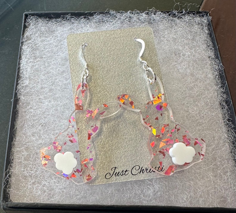 May include: A pair of pink and orange glitter bunny rabbit earrings with white accents. The earrings are displayed in a white box with the text "Just Christi" on the bottom.