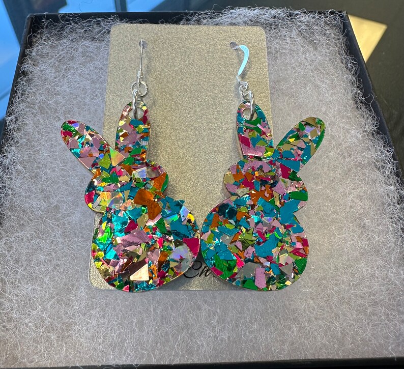 Może przedstawiać: Para kolorowych kolczyk&oacute;w w kształcie kr&oacute;lika wykonanych z mieniącego się brokatu. Kolczyki są srebrne i mają zwisający wz&oacute;r.