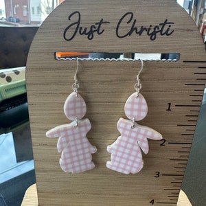 Puede incluir: Un par de pendientes de conejito en forma de gingham rosa y blanco en un expositor de madera con el texto "Just Christi" en la parte superior.