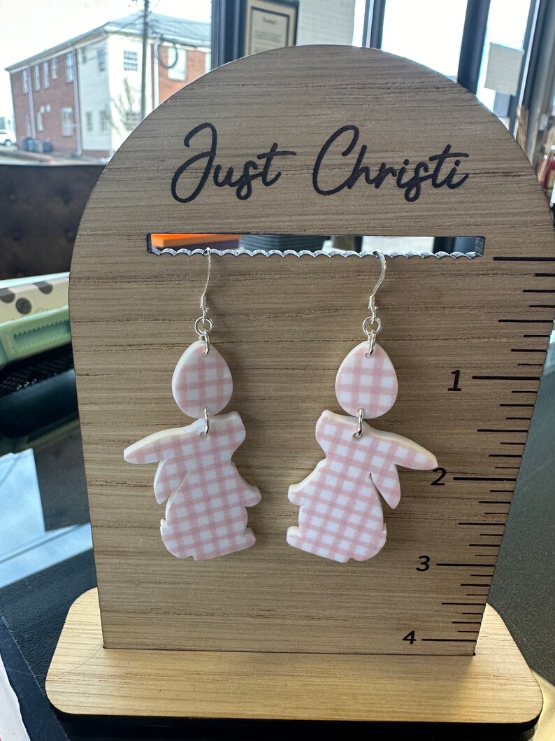 Puede incluir: Un par de pendientes de conejito en forma de gingham rosa y blanco en un expositor de madera con el texto "Just Christi" en la parte superior.