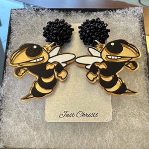 May include: A pair of gold-tone earrings featuring a cartoon hornet design with black and yellow accents. The earrings have black bead dangles.