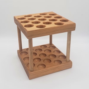 May include: A wooden display stand with two levels, each featuring multiple circular holes. The stand is constructed from light-colored wood and supported by four cylindrical legs. The design is simple and functional, suitable for displaying small items.