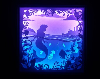 May include: A layered paper art piece depicting an underwater scene with a mermaid, fish, and a castle. The artwork is illuminated with blue and purple light, creating a silhouette effect. The scene includes a mermaid, dolphins, and a castle.