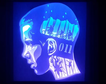 May include: A blue and white layered art piece featuring a profile of a person's face with the number "011" and silhouettes of figures on bicycles. The background depicts a forest scene with trees and a starry sky.