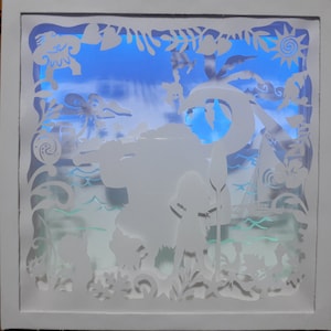 May include: A white, layered paper art piece depicting a tropical scene with a blue sky background. The artwork features silhouettes of characters, palm trees, a boat, and ocean waves, all within a white frame. The scene is illuminated with a blue light.