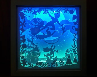 May include: A framed, illuminated shadow box featuring a silhouette scene of Peter Pan, Wendy, John, and Michael flying over Neverland. The artwork includes a pirate ship, moon, stars, and tropical foliage, all in shades of blue and teal.