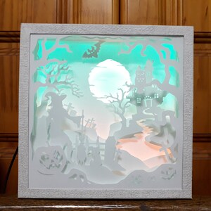 K&ouml;nnte beinhalten: Eine wei&szlig;e, beleuchtete Halloween-Schattenbox. Das geschichtete Design zeigt eine Hexe, ein Spukhaus, einen Vollmond und K&uuml;rbislaternen. Der Hintergrund leuchtet mit teal- und orangefarbenem Licht und erzeugt eine gruselige Atmosph&auml;re. Der Rahmen ist wei&szlig;.