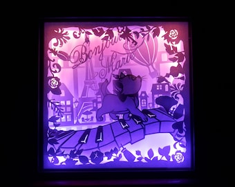 May include: A lit shadow box featuring a cartoon cat playing a piano in front of a Parisian cityscape. The box is illuminated with purple and pink hues, and the words "Bonjour Marie" are written in cursive.