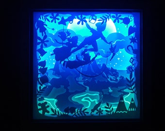 May include: A framed, illuminated shadow box featuring a silhouette scene from Peter Pan. The artwork depicts Peter Pan, Wendy, John, Michael, Tinkerbell, and Captain Hook's ship, all set against a blue and teal backdrop with a full moon.