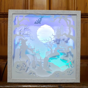 K&ouml;nnte beinhalten: Wei&szlig; gerahmte Halloween-Schattenbox mit einer Hexe, einem Spukhaus und K&uuml;rbissen. Die Szene wird von blauen, violetten und gr&uuml;nen Lichtern beleuchtet, wodurch eine gruselige Atmosph&auml;re entsteht. Ein Vollmond und eine Fledermaus-Silhouette sind ebenfalls zu sehen.