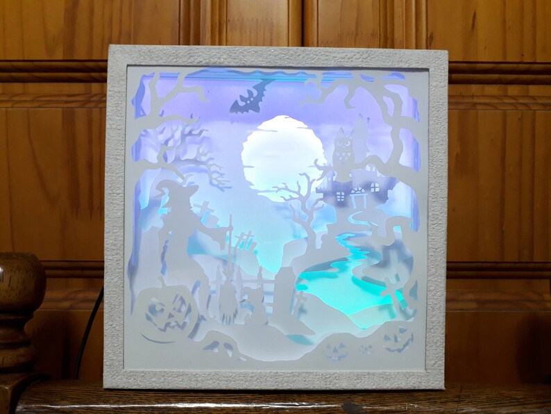 K&ouml;nnte beinhalten: Wei&szlig; gerahmte Halloween-Schattenbox mit einer Hexe, einem Spukhaus und K&uuml;rbissen. Die Szene wird von blauen, violetten und gr&uuml;nen Lichtern beleuchtet, wodurch eine gruselige Atmosph&auml;re entsteht. Ein Vollmond und eine Fledermaus-Silhouette sind ebenfalls zu sehen.