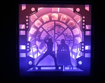 May include: A backlit shadow box featuring a scene from Star Wars. The artwork depicts a lightsaber duel between Luke Skywalker and Darth Vader, set against a backdrop of space and the Millennium Falcon. The color scheme is a gradient of purple and pink.