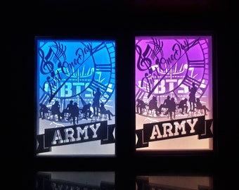 May include: Two illuminated wall art pieces with a clock design, musical notes, and the text "Just One Day", "BTS", and "ARMY". One is lit with blue, the other with purple. Silhouettes of people are visible.