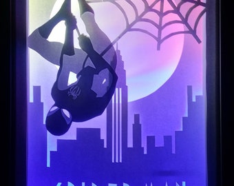 May include: A framed art piece featuring Spider-Man hanging from a web. The artwork has a retro design with a cityscape silhouette, a full moon, and the word "SPIDER-MAN" in stylized text. The color palette includes shades of blue, purple, and white.