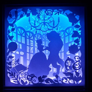 May include: A blue illuminated paper cut light box depicting a dancing couple in silhouette. The design includes a chandelier, roses, and other decorative elements, creating a fairytale scene. The overall effect is a magical ambiance.