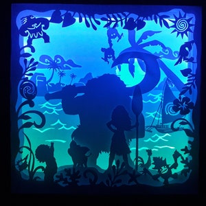May include: A layered paper art piece featuring silhouettes of characters and scenes from a popular animated movie. The artwork depicts a tropical island setting with palm trees, ocean waves, and a sailboat, illuminated with blue and green light.