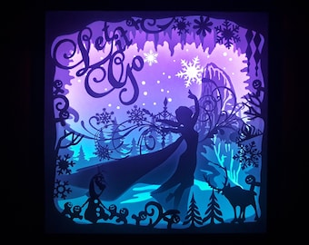 May include: A layered paper art piece with a winter theme. The artwork features a silhouette of a character with outstretched arms, snowflakes, and the words "Let it Go." The color palette includes shades of purple, blue, and white, with a dark background.