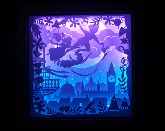 May include: A layered paper art piece depicting Peter Pan and friends flying over London at night. The scene is illuminated with blue and purple hues, featuring silhouettes of the characters, Big Ben, and other iconic landmarks. The artwork is framed in a black box.