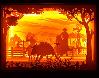 May include: A layered paper art piece depicting a cowboy roping a bull at sunset. The scene is illuminated with warm orange and yellow light, featuring silhouettes of trees, a windmill, and a fence. The artwork evokes a Western theme.