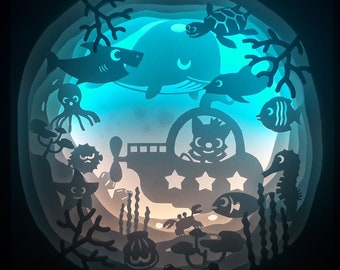 May include: A layered paper art piece depicting an underwater scene. Silhouettes of marine life, including a whale, shark, and seahorse, are layered to create depth. A submarine with a cartoon character is in the center, with a blue and orange gradient background.