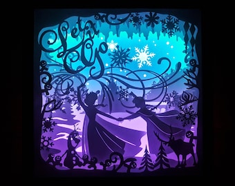 May include: A layered paper art piece featuring a winter scene with the text "Let it Go". Silhouettes of two figures, snowflakes, trees, and a reindeer are visible against a gradient of blue and purple hues. The artwork is illuminated from behind.