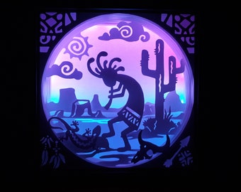 May include: A layered paper art piece depicting a desert scene with a Kokopelli figure playing a flute, a lizard, and a cactus. The artwork is illuminated with purple and blue lighting, creating a vibrant and artistic display.