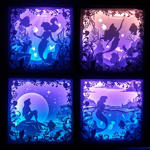 Könnte beinhalten: Vier beleuchtete Schattenboxen mit Meerjungfrau-Silhouetten in Unterwasserszenen. Jede Box hat ein anderes Design, darunter Meerjungfrauen, Burgen, Fische und ein Vollmond, mit blauer, violetter und orangefarbener Beleuchtung.