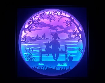 May include: A layered paper art piece depicting a person and a cat fishing on a lake at dusk. The scene is illuminated with blue and purple hues, with trees and a distant shoreline. A bucket sits nearby.