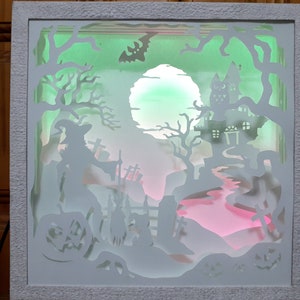 K&ouml;nnte beinhalten: Eine Halloween-Schattenbox mit wei&szlig;em Rahmen. Das geschichtete Design zeigt eine Hexe, ein Spukhaus und einen Friedhof, beleuchtet von gr&uuml;nen, rosa und wei&szlig;en Lichtern. Eine Fledermaus-Silhouette fliegt am Himmel, K&uuml;rbisse sind sichtbar.