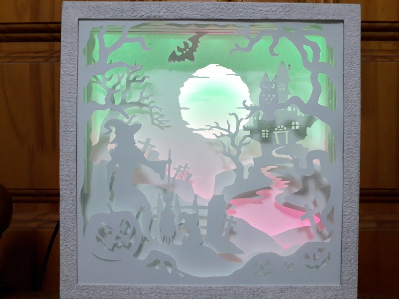 K&ouml;nnte beinhalten: Eine Halloween-Schattenbox mit wei&szlig;em Rahmen. Das geschichtete Design zeigt eine Hexe, ein Spukhaus und einen Friedhof, beleuchtet von gr&uuml;nen, rosa und wei&szlig;en Lichtern. Eine Fledermaus-Silhouette fliegt am Himmel, K&uuml;rbisse sind sichtbar.