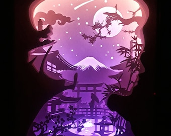 May include: A layered paper art piece depicting a Japanese landscape within a silhouette. The artwork features Mount Fuji, temples, and a full moon, with a color gradient from purple to pink. The silhouette is of a person's profile.