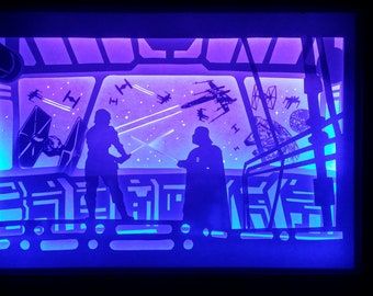 May include: A layered paper art piece depicting a scene from a space opera. Silhouettes of characters with lightsabers face each other in a spaceship interior, with starfighters and a space station visible through the windows. The artwork is illuminated with blue and purple light.