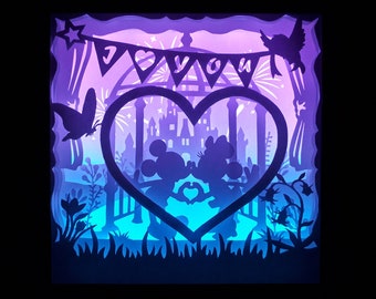 May include: A layered paper art piece with a heart-shaped frame featuring Mickey and Minnie Mouse. The artwork is illuminated with blue and purple hues, with a banner that reads "I LOVE YOU". A butterfly and bird are also included.
