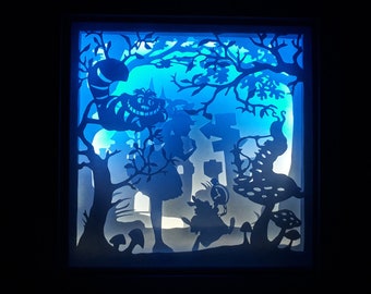 May include: A layered paper art piece depicting a scene from Alice in Wonderland. The artwork features silhouettes of characters like the Cheshire Cat, Alice, and the White Rabbit, set against a blue and white illuminated background, creating a whimsical, fairytale aesthetic.