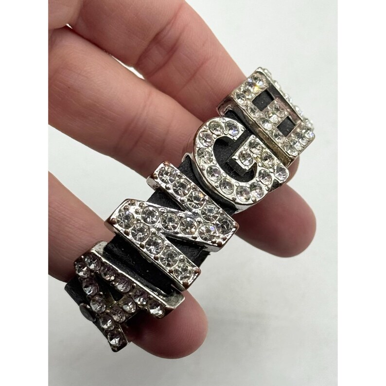 May include: A silver-toned bracelet with the word "MNG" spelled out in large, sparkling, clear rhinestones. The letters are set on a black band. The bracelet is a bold fashion statement.