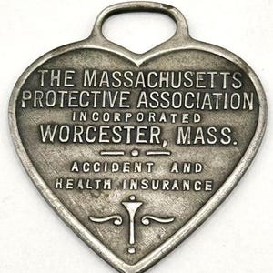 Puede incluir: Un amuleto de plata en forma de corazón con la inscripción "THE MASSACHUSETTS PROTECTIVE ASSOCIATION INCORPORATED WORCESTER, MASS." y "ACCIDENT AND HEALTH INSURANCE".