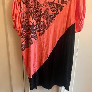 May include: Coral and black asymmetrical top. The coral section features a butterfly pattern in shades of brown and black. The top has short sleeves and a diagonal color block design.